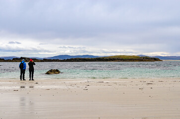 Two people standing on a white sandy beach looking at the sea through binoculars on the Isle of Iona, Scotland.