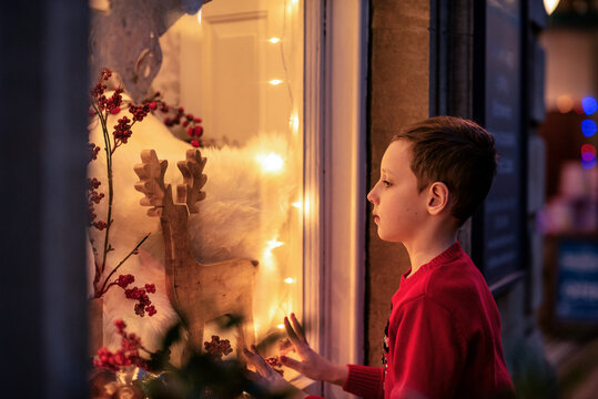 Young boy in red sweater standing outdoors and looking at a festive holiday window display, adorned with lights, decorations, and a reindeer figure, the magic of the season and childhood wonder.