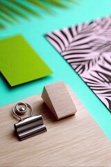 A close-up, overhead shot features office supplies arranged on a bright turquoise surface, including a binder clip, a small wooden block, a green card, and a pi