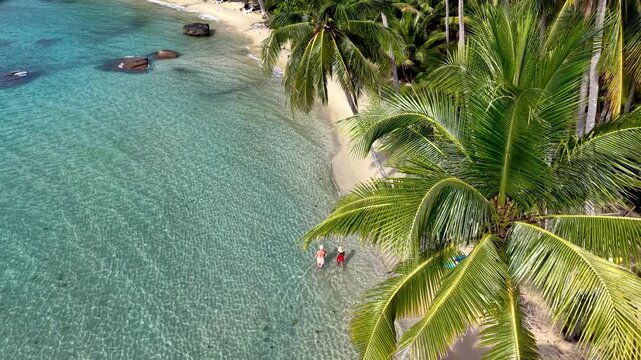 Discover the stunning beaches of Koh Kood Island, Thailand, with crystal clear waters gently lapping at the shore. Lush palm trees provide shade as a couple enjoy a peaceful day by the sea.