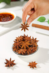 Hand holding star anise spice over bowl of aromatic cooking spices on white kitchen counter
