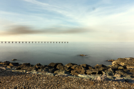 A serene, wide-angle seascape featuring a rough, rocky and gravel shoreline in the foreground, transitioning to the smooth, calm water of the sea. In the mid-ground, a long industrial pier extends int