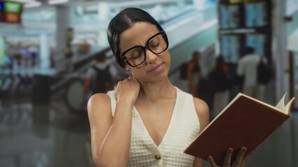 Woman feeling neck pain while reading book in bustling airport terminal, showcasing travel stress with focus on young hispanic woman indoors wearing glasses in a public space.