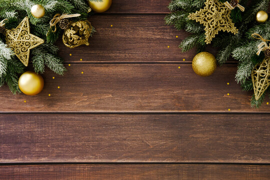 Festive Christmas background with dark wood planks and snow-dusted fir branches framing the top. Adorned with glittering gold stars, balls, and pine cones. Copy space in center.