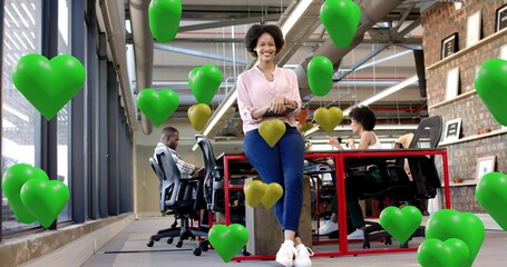 Woman in pink blouse leaning on red desk, open office, holding tablet, green gold heart balloons