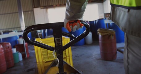 Gripping gloved worker in safety vest using black pallet jack handle in workshop, with yellow crate