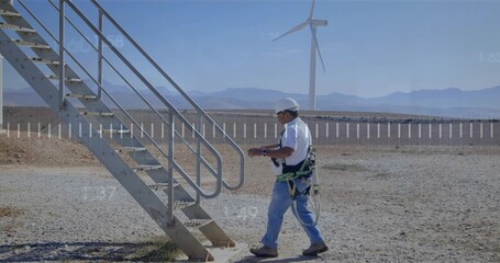 Walking tech wearing hard hat and harness holding device to staircase at wind farm, copy space