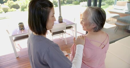 Sitting mother wearing pink strappy top talking with daughter in grey and white on wooden deck