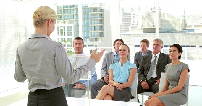 Presenting woman in business wear at clear lectern addressing group in office with glass windows
