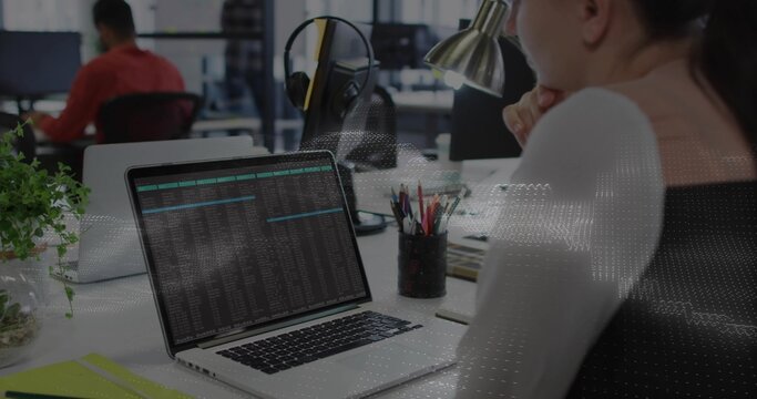 Leaning woman in white top studying laptop showing terminal data at open-plan office, headphones