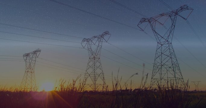 Fototapeta Silhouetting three high-voltage towers and power lines over rural field at sunset with tall grasses