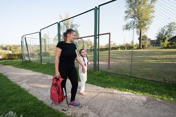 Mother and daughter stroll along a path beside a fenced field, sharing a moment of connection on a sunny day.