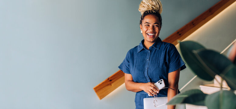 Smiling woman with blonde braided bun holding smartphone and tablet at staircase - Powered by Adobe