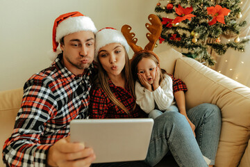 Christmas family wearing Santa hats and reindeer antlers making funny faces while taking a selfie with a tablet, sitting together by a decorated  xmas  tree.