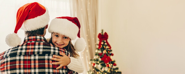 Little girl in Santa hat hugs her father with a joyful smile, both dressed in cozy holiday outfits, with a decorated Christmas tree glowing in the background.