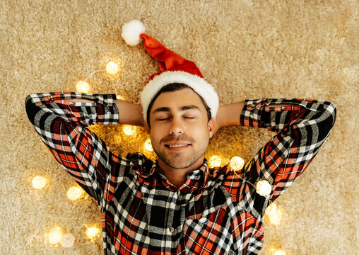 Peaceful man in plaid shirt and Santa hat lying on the carpet with eyes closed, surrounded by glowing lights, dreaming happily about love, New Year joy, and festive Christmas atmosphere.