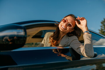 sunset woman adjusting sunglasses through car window smiling, urban skyline background, modern outfit, relaxed posture, reflective