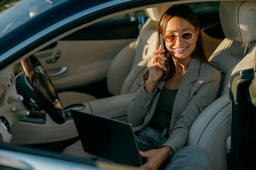 Woman working on laptop inside car, making playful peace sign during video call while managing schedule, blazer-clad professional