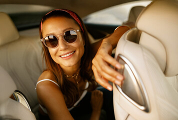 Smiling woman leaning forward in backseat wearing sunglasses and bandana, candid summer travel vibe in leather interior with warm