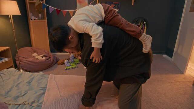 Asian father lifting his little aon on his back, laughing and putting him down on soft bed in warmly lit cozy bedroom during joyful evening family time