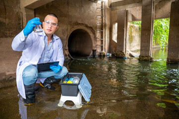 Scientist Taking Environmental Water Sample with plastic glass and Protective Gear Near Drainage Tunnel