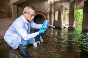 Scientist Taking Environmental Water Sample with plastic glass and Protective Gear Near Drainage Tunnel