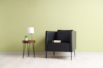 Blurred view of black armchair and coffee table with desk lamp near green wall
