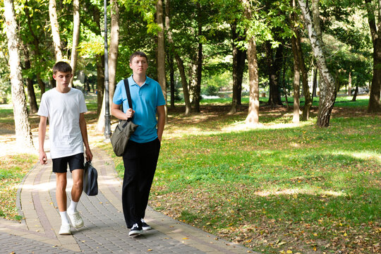 Two figures walk through a sunlit park. Trees towering overhead and shadows dappling the grassy landscape on a lovely summer's day.