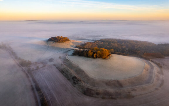 Aerial view of golden light kissing the misty Wittenham Clumps, painting the fields in a soft, ethereal glow, Oxfordshire, England, United Kingdom.