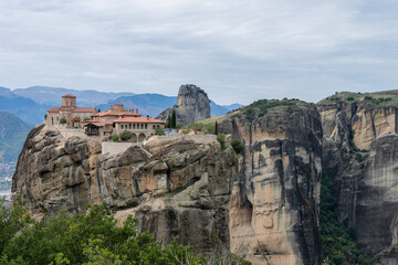 A monastery with red-tiled roofs stands atop a towering sandstone cliff in Meteora, surrounded by lush greenery and distant misty mountains.