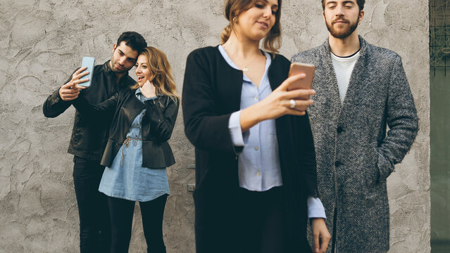 Group of young friends socializing with phones in the city. Woman shows content on her smartphone while another couple takes a selfie in the background, concept of digital connection.