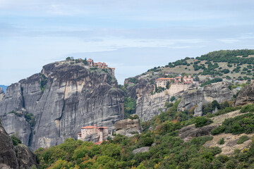 Monasteries crown towering sandstone cliffs in Meteora, surrounded by lush hills and dramatic rock...