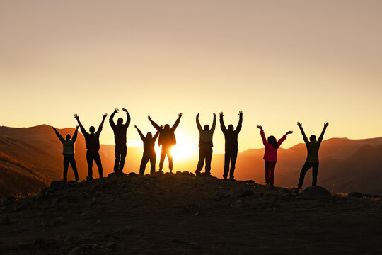 Silhouettes of hikers with raised arms standing on mountain summit at sunset, celebrating success, victory and teamwork