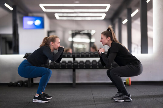 Two women perform squats together in a modern gym during a fitness session focused on strength training and camaraderie