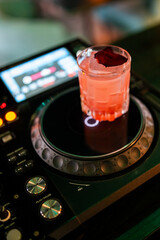 A vibrant pink cocktail, garnished with a dried berry or leaf, sits on a black professional DJ turntable mixer with control buttons and a screen in a dimly lit bar or club setting