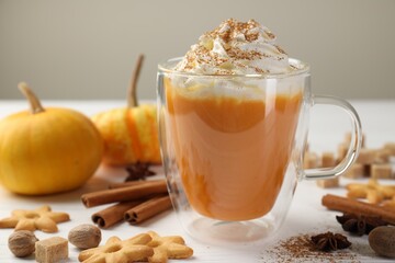 Tasty pumpkin latte with whipped cream in glass cup, cookies and ingredients on white wooden table, closeup