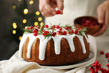 Woman putting cranberries onto tasty Christmas cake with icing and rosemary at table, closeup
