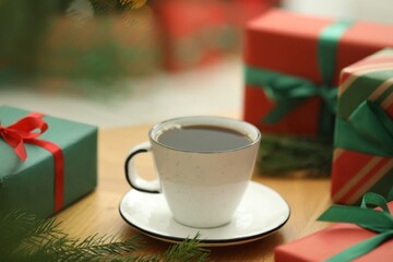 Aromatic coffee in cup, fir tree branches and gift boxes on wooden table against blurred lights, closeup. Christmas greeting card