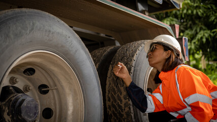 A woman in bright orange safety gear carefully examines the tires of a large truck at a construction site. She focuses on ensuring proper maintenance and safety for the vehicle © Happy Photo