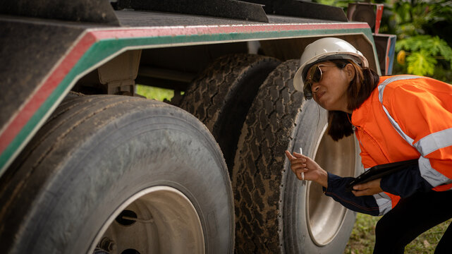 A woman wearing a hard hat and reflective jacket closely examines the tires of a truck at a construction site during daytime, ensuring safety standards - Powered by Adobe