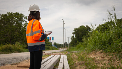A construction worker stands on a pathway, dressed in bright safety gear, using a tablet to check...