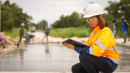 A construction worker in a bright safety jacket and hard hat reviews data on a tablet near workers pouring concrete at a site. It is a sunny day, and the area is active with construction activity