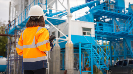 A construction worker in a safety vest and helmet monitors activity on a construction site filled with blue machinery in an urban setting. The sky is clear, indicating a sunny day