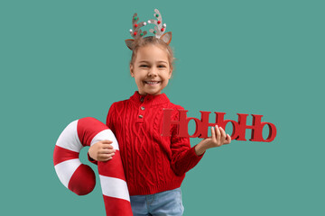 Happy little girl in Christmas reindeer horns with toy candy cane and word HOHOHO on green background