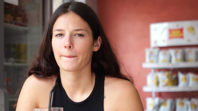Thoughtful Woman with long dark hair in a black sleeveless top holds a drink with a straw at a cafe, shelves with packaged goods in the background.