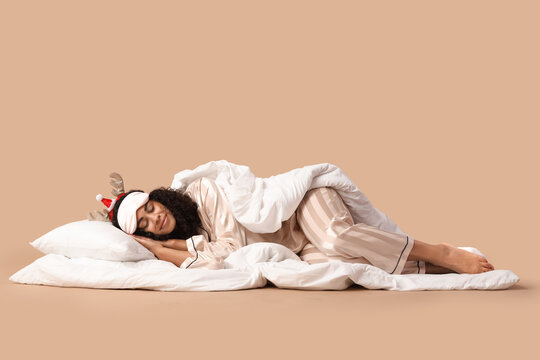 Pretty young African-American woman in pajamas and reindeer horns sleeping on beige background. Christmas celebration
