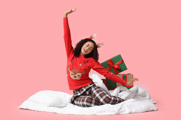 Pretty young African-American woman in pajamas and reindeer horns stretching on pink background. Christmas celebration