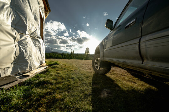 An off-road vehicle with mud tires stands on a green meadow near a hut during golden hour. A green forest and snow-dusted mountains are visible in the background.