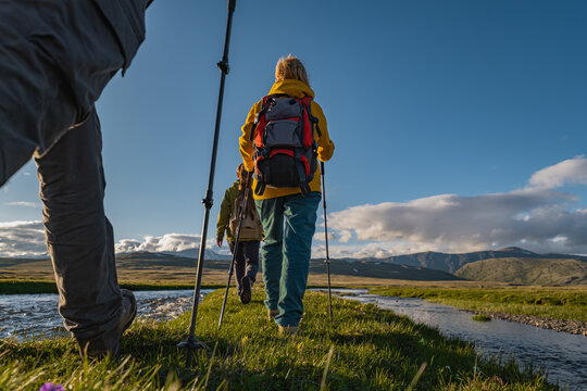 A group of hikers in bright clothing with backpacks and trekking poles walks across a green meadow flanked by rivers. Golden hour light illuminates the mountain backdrop and blue sky.