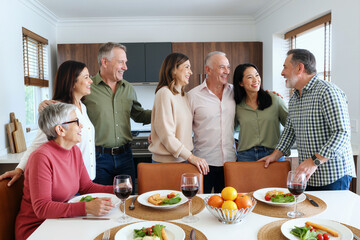 Group of middle aged and senior multiethnic men and women standing together in modern kitchen smiling and talking before meal, plates of food and glasses of wine on table